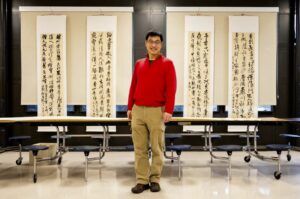 Dr Jun Lin smiles in front of four hanging calligraphy pieces